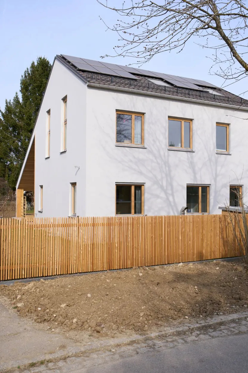Weißes Giebelhaus mit Holzfenstern und Photovoltaik auf dem Dach, davor ein Holzlattenzaun, Eckansicht von der Straße