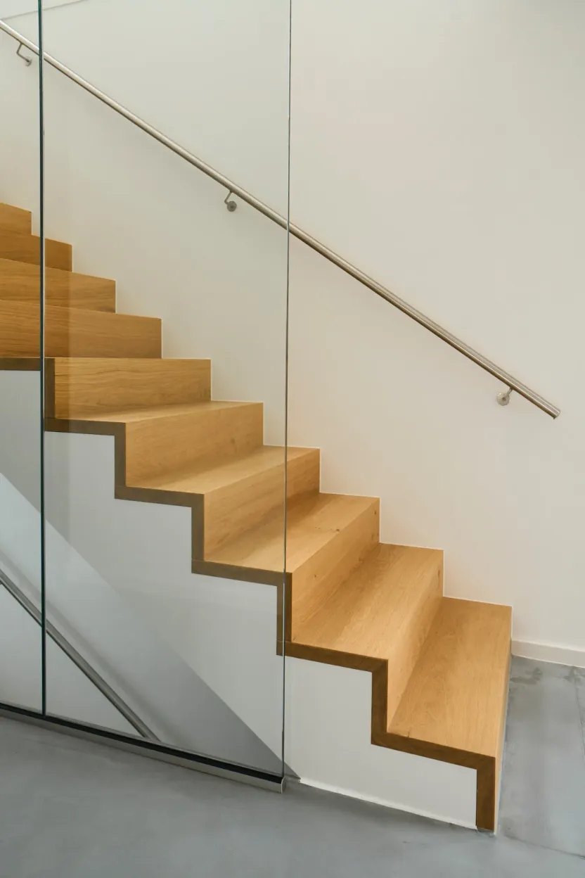 Holztreppe vor weißer Wand mit Edelstahlhandlauf, im Vordergrund eine Glaswand