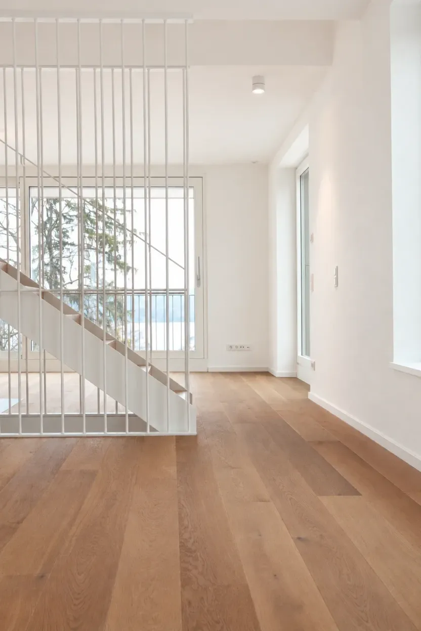 Wohnraum mit Holzboden, Treppe hinter weißem Stabgeländer, dahinter Fenstertür mit Blick auf Bäume und den See