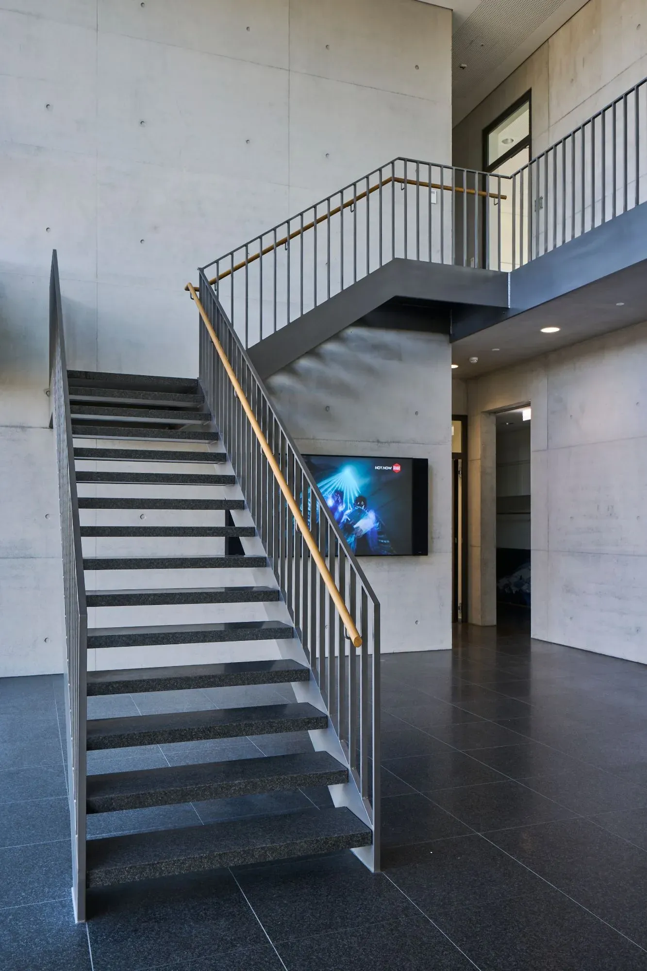 Foyer mit Stahltreppe, Granitstufen und Holzhandlauf vor Sichtbetonwand