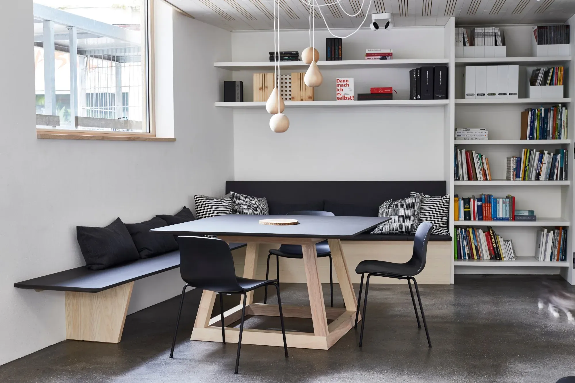 Eckbank mit schwarzen Kissen, Holztisch und schwarzen Stühlen vor weißem Bücherregal