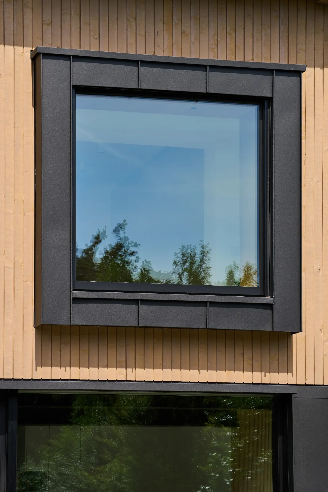 Quadratisches Fenster mit dunkler Metalleinfassung in der senkrechten Holzschalung, darunter die Glasfront