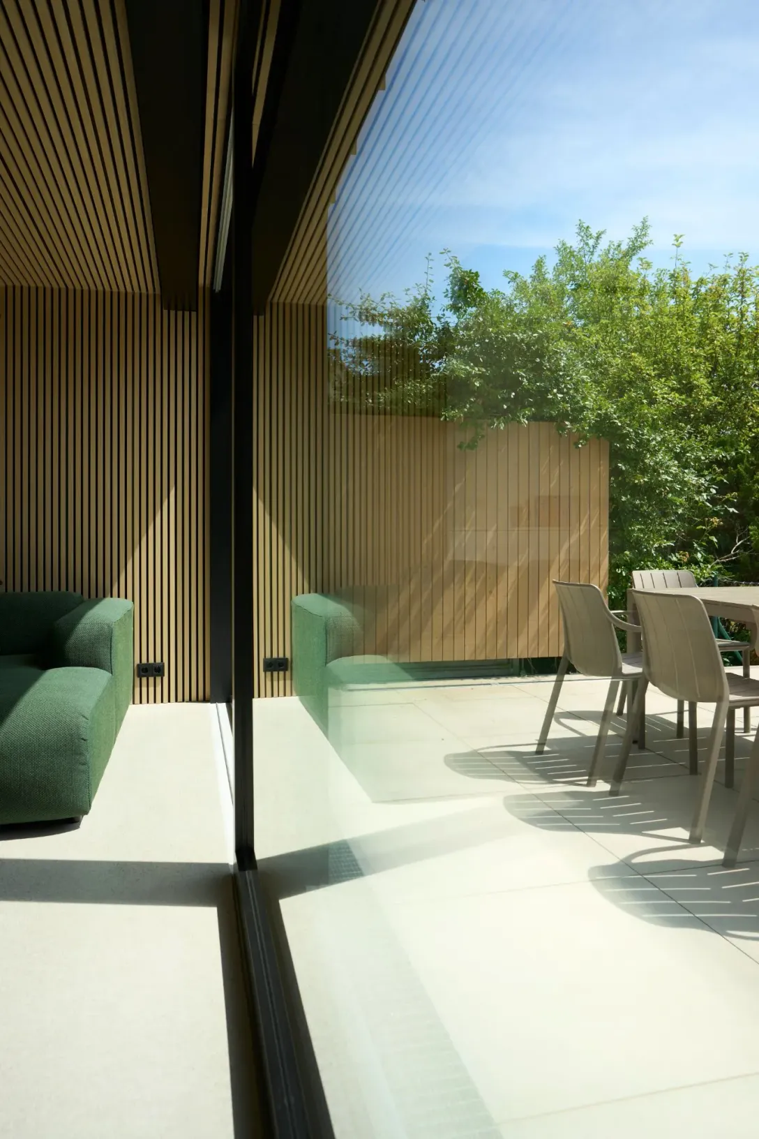 Glasfront zwischen Wohnraum mit grünem Sofa und Holzlamellenwand und der Terrasse mit Gartenstühlen