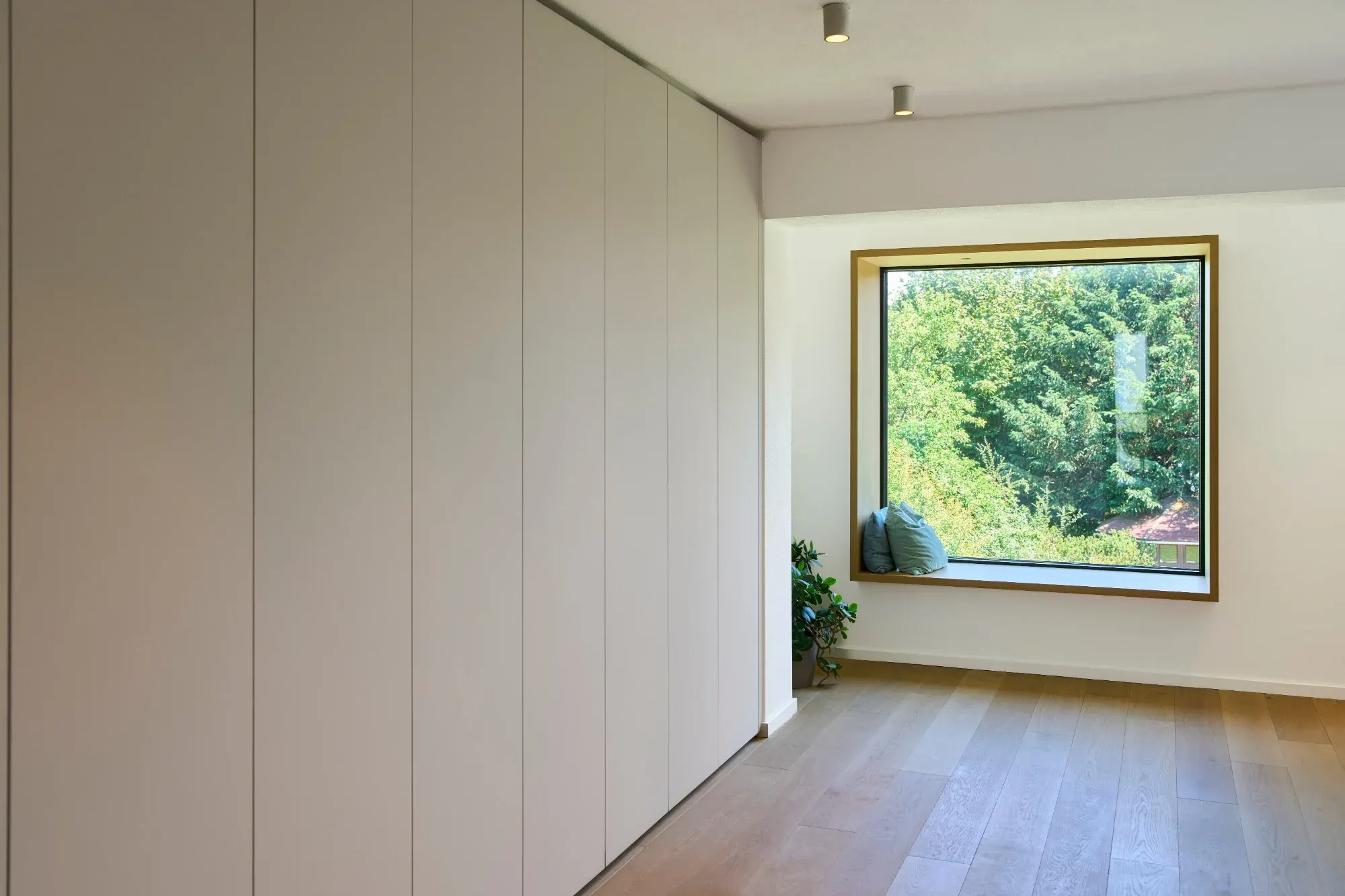 Raum mit weißer, grifflos durchlaufender Schrankwand und tiefem Sitzfenster mit Holzrahmen und Blick ins Grüne
